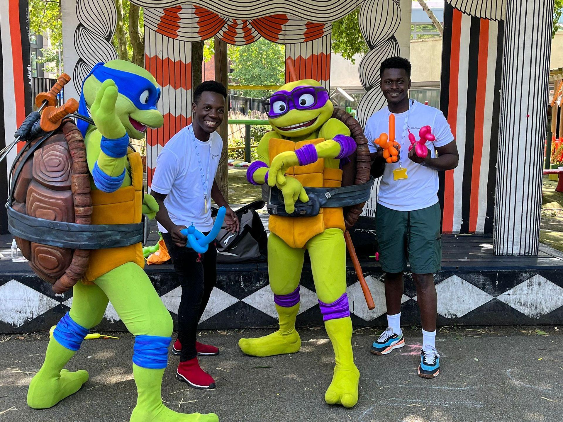 Two people standing with characters dressed as turtles holding balloon animals at an outdoor event.