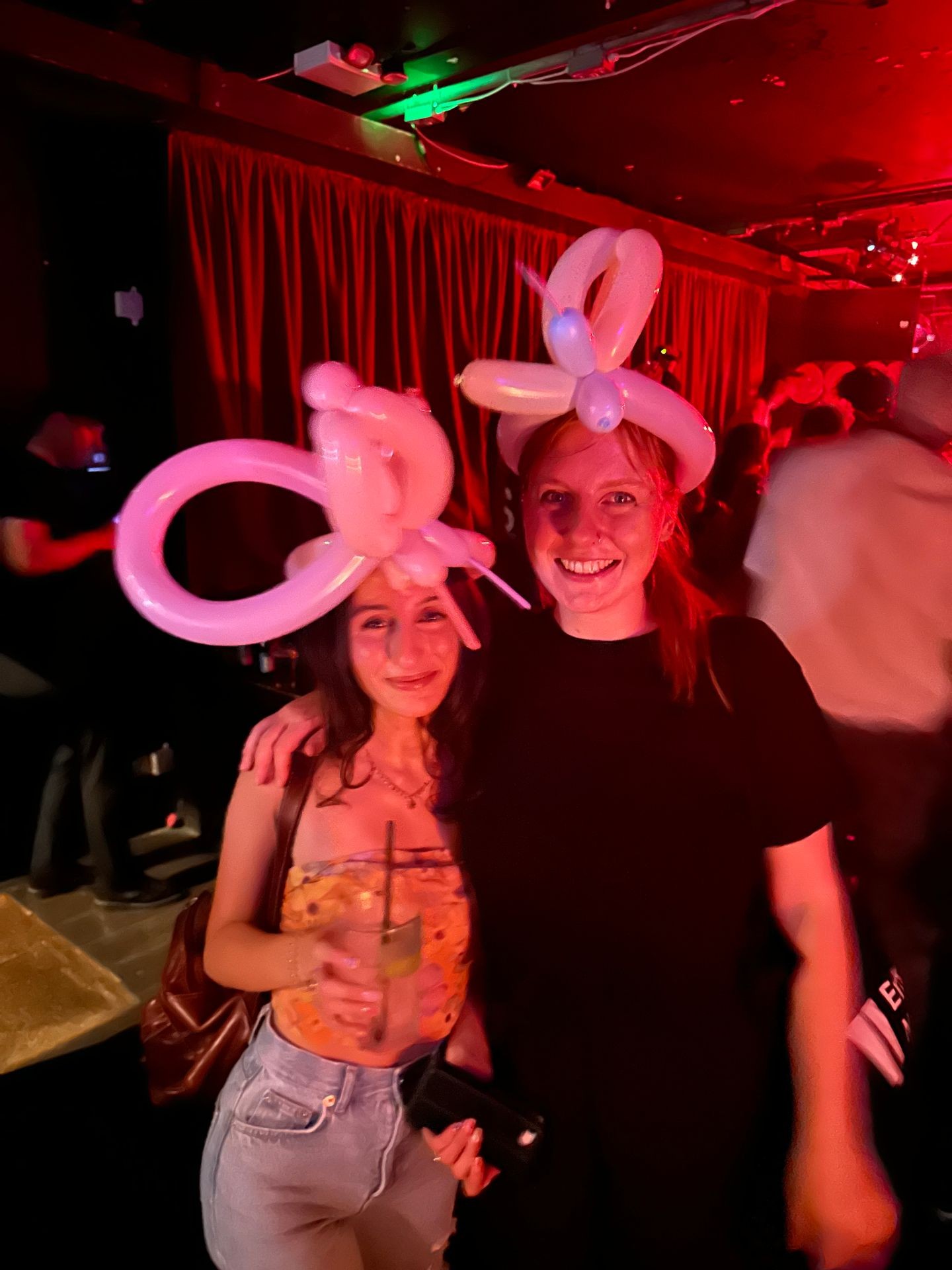 Two people wearing balloon hats at a party with dim red lighting and a background curtain.