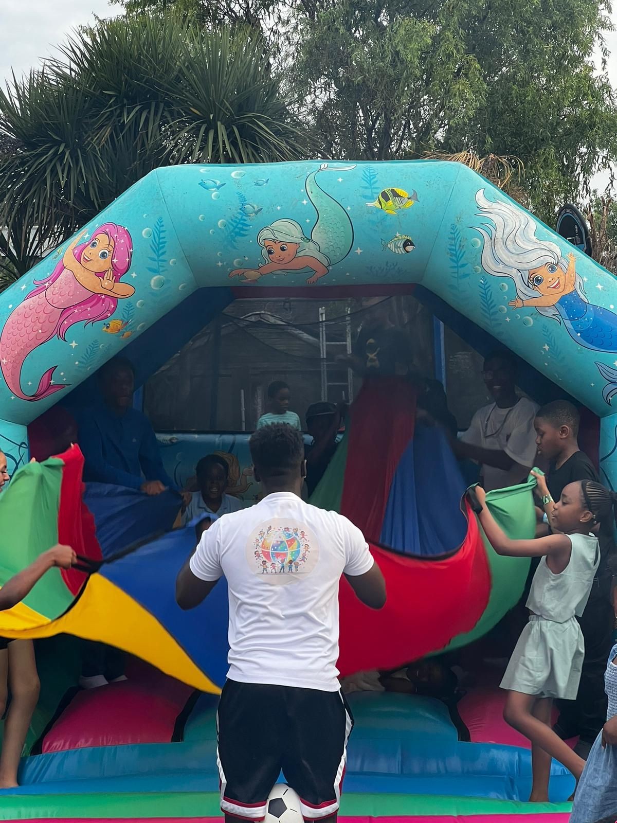 Children playing with colorful fabric in front of an inflatable bouncy house with mermaid decorations outdoors.